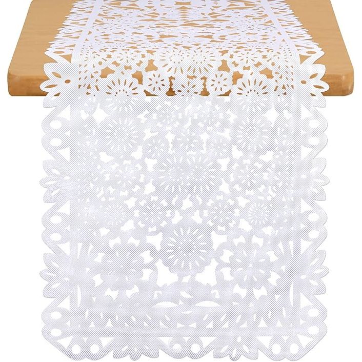 This Mexican party table runner measures 14 x 84 inches and features traditional white papel picado designs. Papel picado is a decorative craft made by cutting intricate patterns into paper or fabric, often used for celebrations and fiestas in Mexican culture.